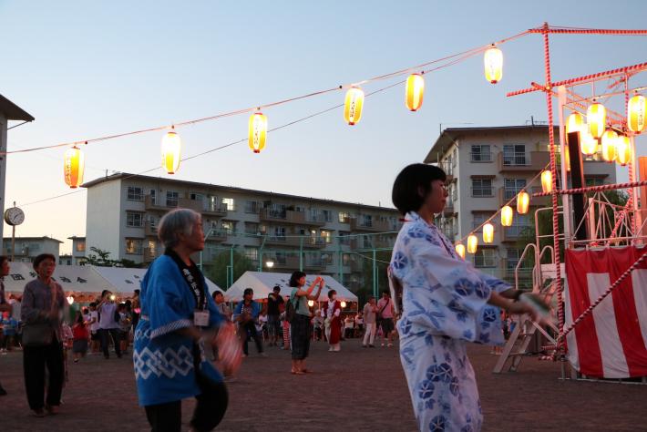 紅白幕のかかったやぐらの周りで法被やゆかたをきて踊る市民。頭上にはちょうちん、背景に団地が数棟立っている。