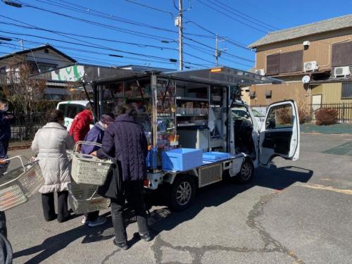 商品をたくさん積んだ車で買い物をする人たちがかごを持って並んでいる様子